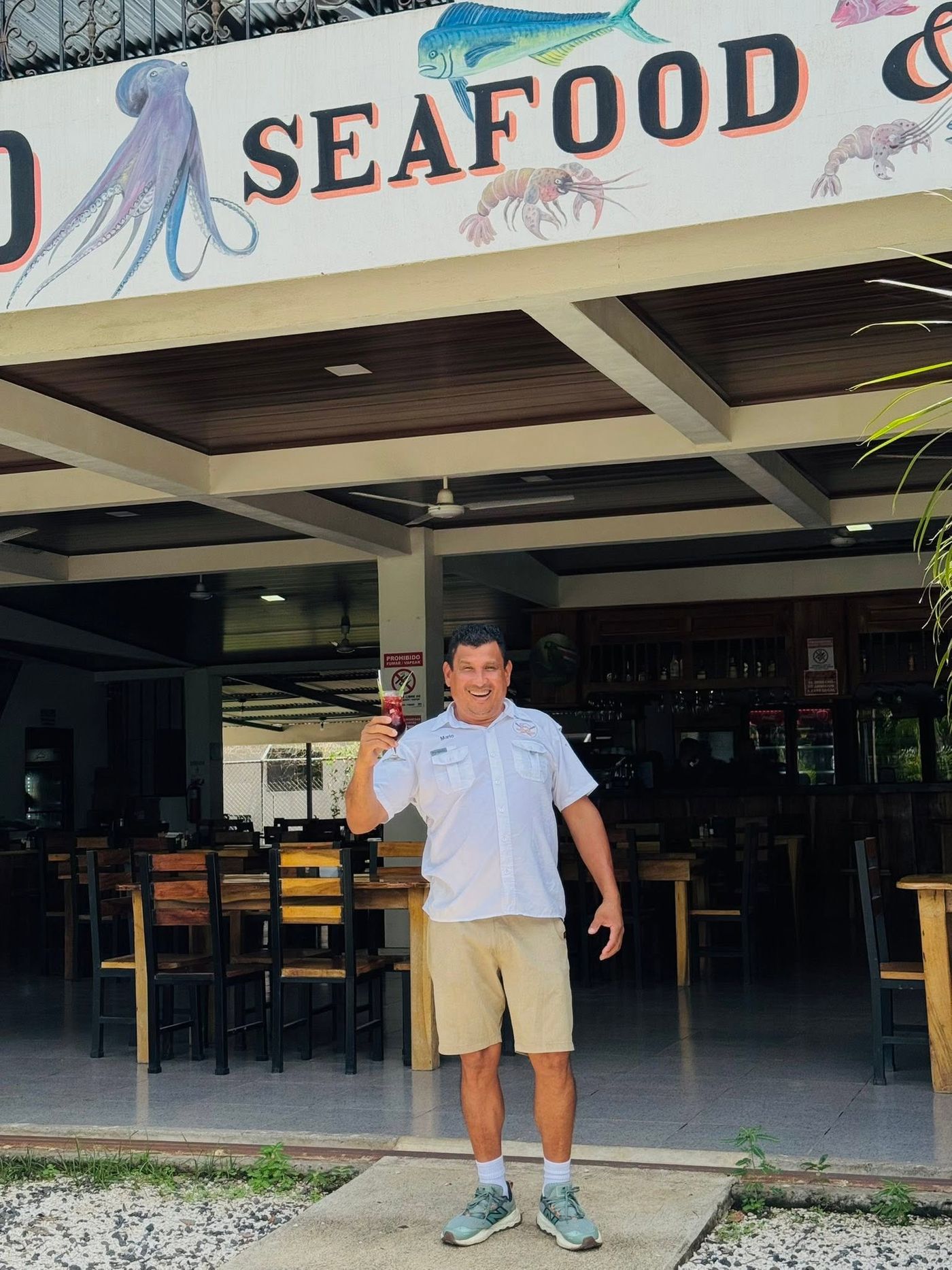 Mario brinda con una copa de sangría frente al restaurante, bajo el letrero pintado de mariscos